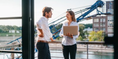 Two business professionals standing on a balcony, engaged in a thoughtful discussion, with a city skyline in the background.