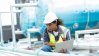 Technician in a hard hat inspecting outdoor HVAC equipment with a laptop.