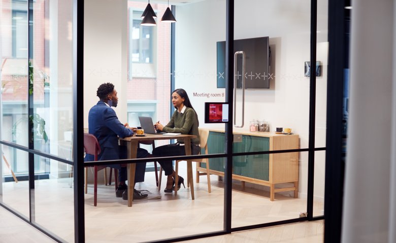 Two people in a meeting room inside a workspace with glass walls..