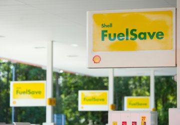 Shell FuelSave signage at a gas station with yellow signs and fuel pumps in the background.