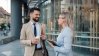 Two business professionals engaged in a casual conversation, standing in a modern office environment.