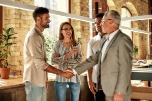 Two business people shaking hands and smiling while standing with coworkers in the creative office.