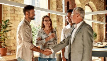 Two business people shaking hands and smiling while standing with coworkers in the creative office.
