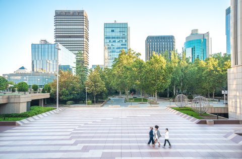 Tall real estate buildings in the back with persons walking through a park 