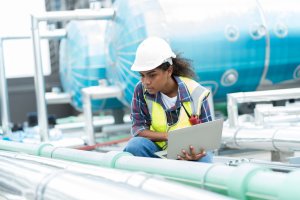 A maintenance technician inspects industrial piping with a laptop, wearing safety gear, in front of blue machinery.