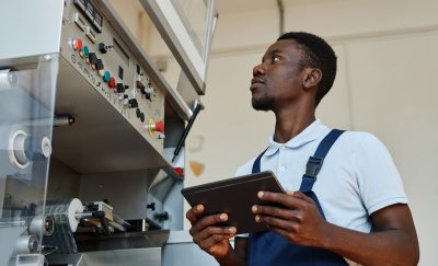 Factory worker inspecting machinery while holding a tablet for digital maintenance checks.