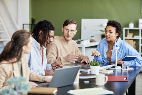 Group of diverse colleagues collaborating around a table in a modern office, discussing work with laptops, notebooks, and coffee.