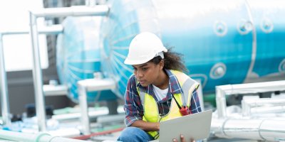 Technician in a hard hat inspecting outdoor HVAC equipment with a laptop.