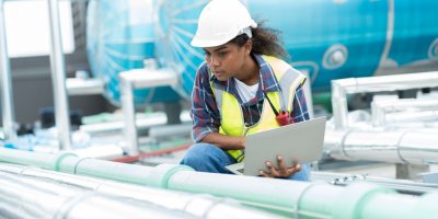 A maintenance technician inspects industrial piping with a laptop, wearing safety gear, in front of blue machinery.