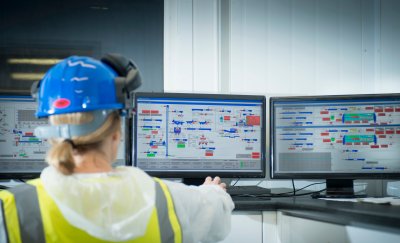 Maintenance woman in blue hard hat and high visibility yellow vest pointing at screens with data in front of her.