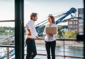 Two business professionals standing on a balcony, engaged in a thoughtful discussion, with a city skyline in the background.