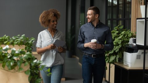 Two colleagues walking through an office, smiling and talking while holding a coffee and a laptop.