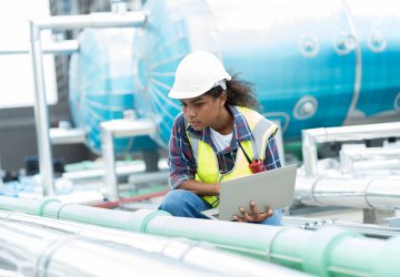 Technician in a hard hat inspecting outdoor HVAC equipment with a laptop.