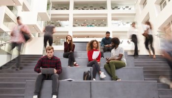 Higher education students sitting on the stairs in the hall of a building.