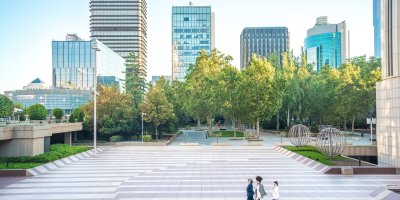 Tall real estate buildings in the back with persons walking through a park.