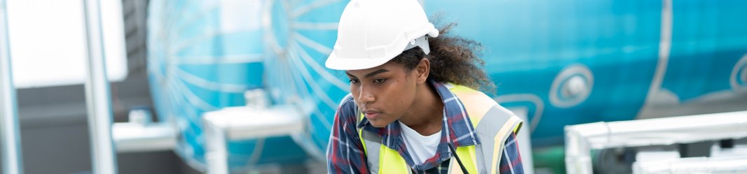 Technician in a hard hat inspecting outdoor HVAC equipment with a laptop.