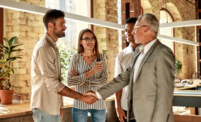 Two business people shaking hands and smiling while standing with coworkers in the creative office.
