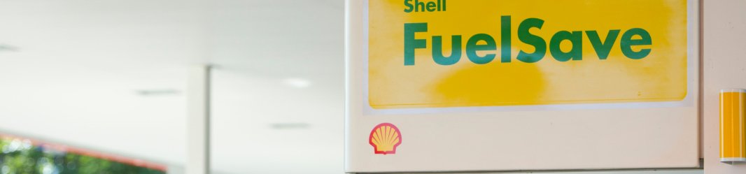 Shell FuelSave signage at a gas station with yellow signs and fuel pumps in the background.