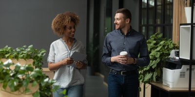 Two colleagues walking through an office, smiling and talking while holding a coffee and a laptop.