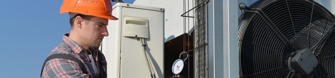 A worker using a pressure gauge to inspect an HVAC system, wearing a hard hat and overalls.