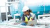 Technician in a hard hat inspecting outdoor HVAC equipment with a laptop.