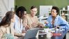 Group of diverse colleagues collaborating around a table in a modern office, discussing work with laptops, notebooks, and coffee.