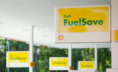 Shell FuelSave signage at a gas station with yellow signs and fuel pumps in the background.