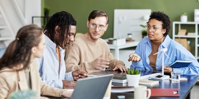 Group of diverse colleagues collaborating around a table in a modern office, discussing work with laptops, notebooks, and coffee.