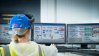 Maintenance woman in blue hard hat and high visibility yellow vest pointing at screens with data in front of her.