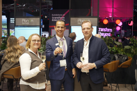 Three attendees holding glasses and posing together at the Planon Real Estate stand.