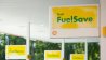 Shell FuelSave signage at a gas station with yellow signs and fuel pumps in the background.