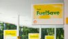 Shell FuelSave signage at a gas station with yellow signs and fuel pumps in the background.