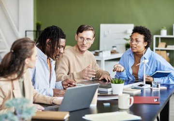 Group of diverse colleagues collaborating around a table in a modern office, discussing work with laptops, notebooks, and coffee.
