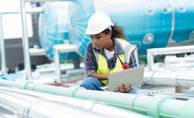 A maintenance technician inspects industrial piping with a laptop, wearing safety gear, in front of blue machinery.