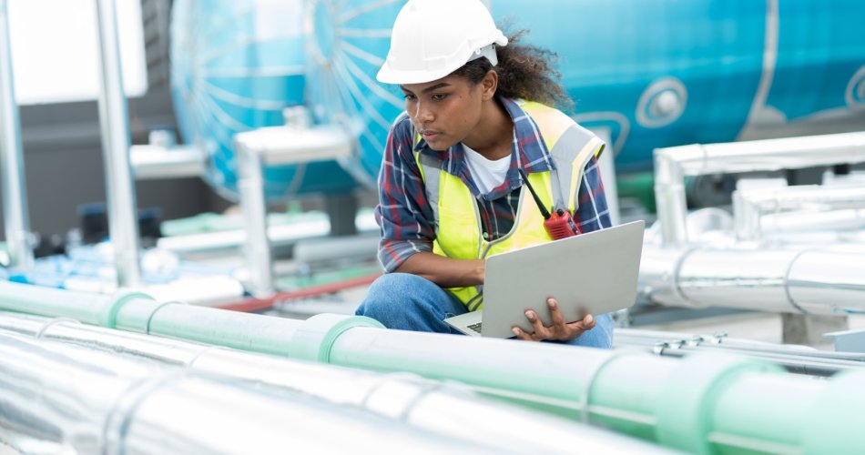 A maintenance technician inspects industrial piping with a laptop, wearing safety gear, in front of blue machinery.