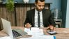 A man in a black suit working on financial data with a calculator.