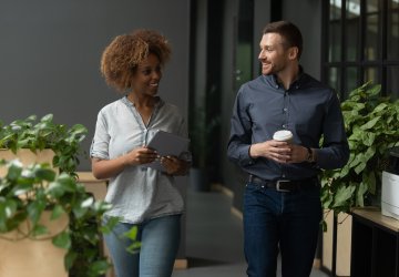 Two colleagues walking through an office, smiling and talking while holding a coffee and a laptop.