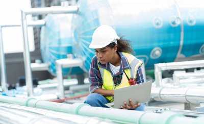 Technician in a hard hat inspecting outdoor HVAC equipment with a laptop.