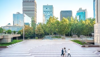 Tall real estate buildings in the back with persons walking through a park.