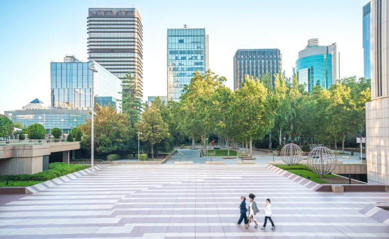 Tall real estate buildings in the back with persons walking through a park.