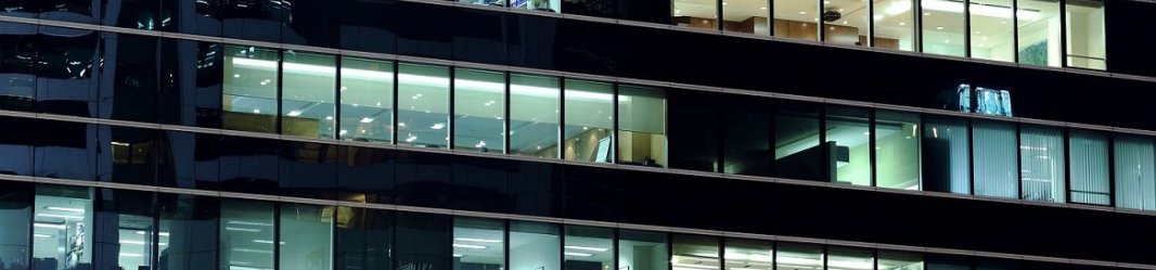Nighttime view of an office building with illuminated workspaces visible through large windows.